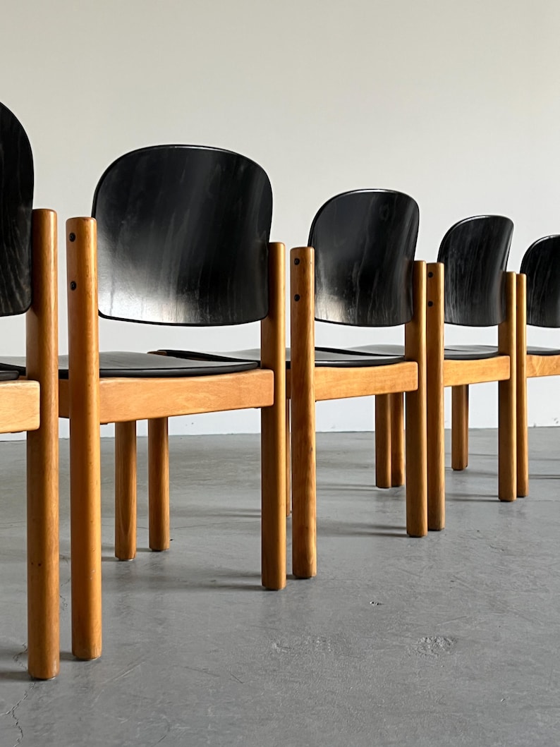 May include: A row of mid-century modern chairs. Each chair has a black, curved back and seat, supported by a light-colored wooden frame and legs. The chairs are arranged in a line against a neutral background.