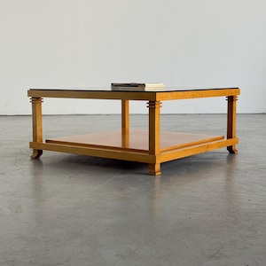 May include: A square coffee table constructed from light-toned wood featuring a dark glass top surface and a lower shelf. The table has four straight legs with decorative tiered detailing at the top and small feet at the base of each leg.