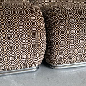 May include: Close-up of a set of upholstered furniture with a geometric pattern. The fabric features a repeating pattern of small squares in shades of brown, beige, and black. The furniture has a low profile and rests on a silver metal base.