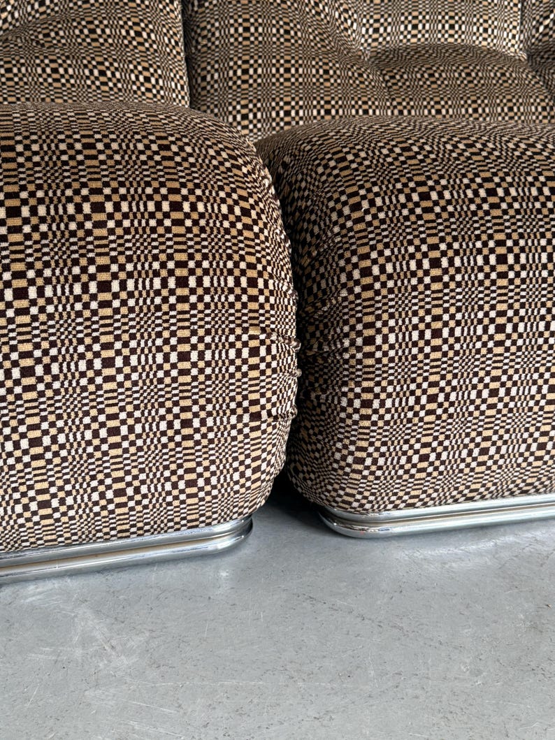 May include: Close-up of a set of upholstered furniture with a geometric pattern. The fabric features a repeating pattern of small squares in shades of brown, beige, and black. The furniture has a low profile and rests on a silver metal base.