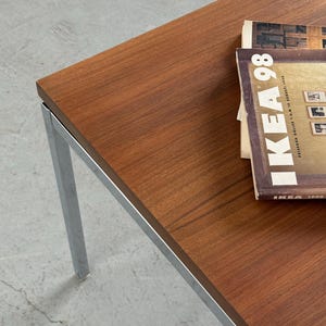 Op de afbeelding: Een rechthoekige salontafel met een blad in houtnerf en chromen poten. Op de tafel ligt een stapel tijdschriften met de tekst "IKEA 98" zichtbaar. Afmetingen: ongeveer 120 cm lang en 60 cm breed.