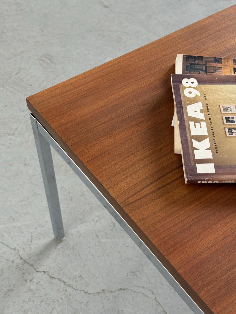 Op de afbeelding: Een rechthoekige salontafel met een blad in houtnerf en chromen poten. Op de tafel ligt een stapel tijdschriften met de tekst "IKEA 98" zichtbaar. Afmetingen: ongeveer 120 cm lang en 60 cm breed.