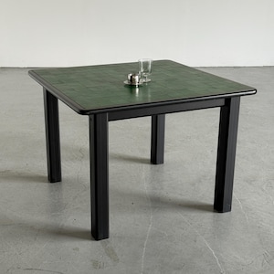 Pode incluir: Uma mesa quadrada com um tampo de azulejos verde-escuro e uma estrutura preta com quatro pernas robustas. Uma pequena chávena de metal prateado sobre um pires a condizer e um copo de vidro transparente estão colocados no centro da mesa.