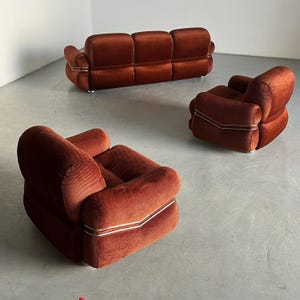 May include: A three-piece burnt orange velvet living room set, including a sofa and two armchairs. The furniture features a distinctive mid-century modern design with sleek, metallic accents. The upholstery is a rich, textured velvet in a warm burnt orange hue. The sofa has three cushioned seats, and the armchairs are similarly styled with rounded forms and padded armrests. The metallic trim adds a touch of contrast and sophistication to the overall design. The set is ideal for adding a vintage vibe to a contemporary living space.