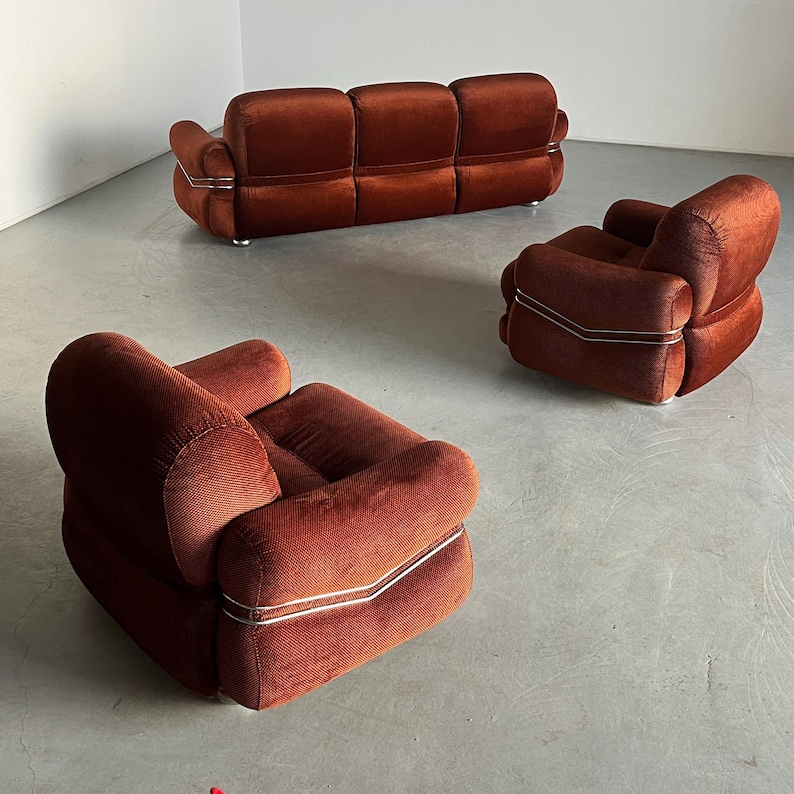 May include: A three-piece burnt orange velvet living room set, including a sofa and two armchairs. The furniture features a distinctive mid-century modern design with sleek, metallic accents. The upholstery is a rich, textured velvet in a warm burnt orange hue. The sofa has three cushioned seats, and the armchairs are similarly styled with rounded forms and padded armrests. The metallic trim adds a touch of contrast and sophistication to the overall design. The set is ideal for adding a vintage vibe to a contemporary living space.