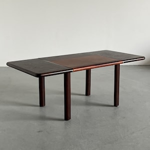 May include: A dark brown wooden dining table with a rectangular top and rounded corners. The table has four cylindrical legs and a center section of a lighter brown colour. The table is approximately 152 cm long.