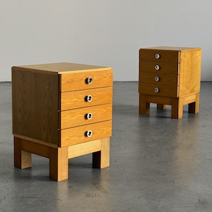 May include: Two light brown wooden bedside tables, each featuring four stacked drawers with circular silver-toned metal pulls. The cabinets are supported by four thick, block-style legs and display a visible wood grain pattern throughout the exterior.
