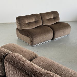 May include: Two-piece brown patterned sofa with a low profile and rounded edges. The sofa features a geometric design and a tufted backrest. The base has a silver metal trim. The sofa is set against a plain white wall and a gray floor.