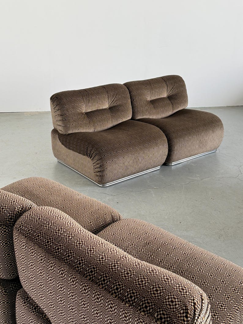 May include: Two-piece brown patterned sofa with a low profile and rounded edges. The sofa features a geometric design and a tufted backrest. The base has a silver metal trim. The sofa is set against a plain white wall and a gray floor.