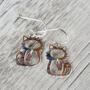 May include: A pair of silver cat-shaped dangle earrings with a small charm hanging from each cat.