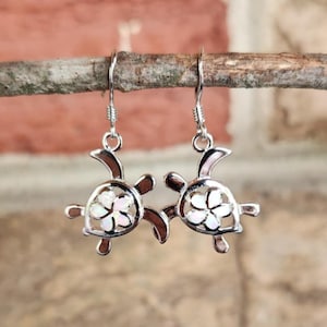 May include: A pair of silver-toned turtle earrings with a flower design in the center of the shell. The earrings are hanging from a small branch against a brick wall background. The turtle's flippers and head are visible.