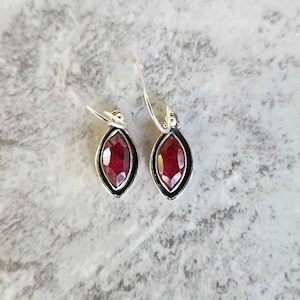 May include: A pair of silver hoop earrings with a red gemstone pendant. The earrings are simple and elegant.