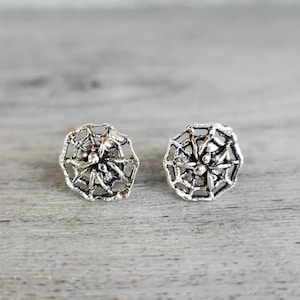 May include: Two silver-colored stud earrings with a spiderweb design. The earrings are round and have a detailed, openwork pattern. They are displayed on a light gray wooden surface.