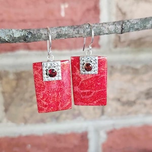 May include: A pair of red rectangular earrings with a silver square top and a small red gemstone in the center. The earrings are hanging from a silver hook. The earrings are displayed on a small branch.