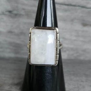 May include: A silver ring featuring a large, rectangular, semi-translucent white moonstone cabochon set in a polished metal frame. The setting has a decorative border with small vertical metallic bars along the sides of the stone.