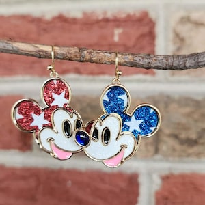 K&ouml;nnte beinhalten: Ein Paar goldfarbene Mickey Mouse-Ohrringe. Ein Ohrring hat roten Glitzer mit wei&szlig;en Sternen, der andere blauen Glitzer mit wei&szlig;en Sternen. Beide haben wei&szlig;e Gesichter, schwarze Augen, rosa M&uuml;nder und einen blauen Edelstein.