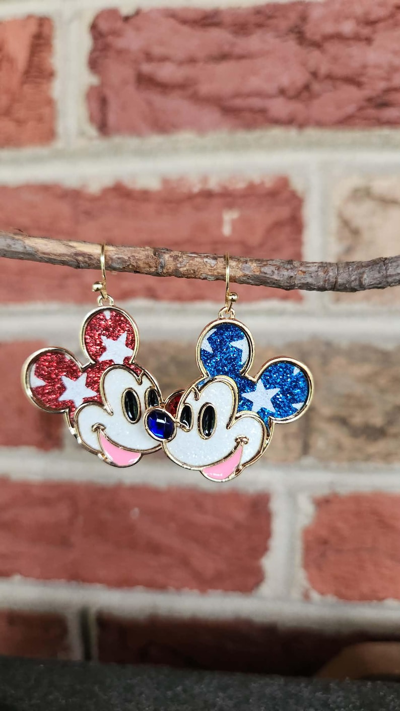 K&ouml;nnte beinhalten: Ein Paar goldfarbene Mickey Mouse-Ohrringe. Ein Ohrring hat roten Glitzer mit wei&szlig;en Sternen, der andere blauen Glitzer mit wei&szlig;en Sternen. Beide haben wei&szlig;e Gesichter, schwarze Augen, rosa M&uuml;nder und einen blauen Edelstein.