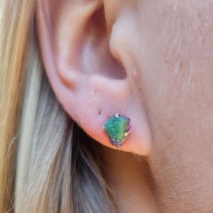 May include: Close-up of a stud earring featuring a raw, uncut gemstone. The earring has a vibrant mix of green and purple hues, with a gold-colored setting. The earring is set against the backdrop of a person's ear and blonde hair.