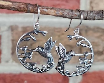 May include: A pair of silver-toned hummingbird earrings. Each earring features a detailed hummingbird within a circular frame with floral accents. The earrings are suspended from simple ear wires, showcasing a nature-inspired design.