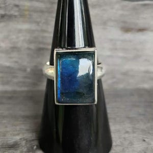 May include: A silver ring featuring a rectangular labradorite gemstone with a polished surface. The stone displays deep blue and dark grey tones with a reflective sheen set within a smooth metal bezel on a simple, plain silver band.