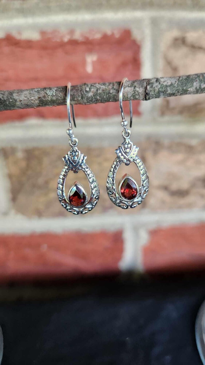 May include: A pair of silver-toned drop earrings. Each earring features a teardrop-shaped frame with a detailed, ornate design. A vibrant, red gemstone is set within the frame. The earrings are suspended from a simple hook.