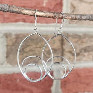 May include: A pair of silver-colored earrings with a modern design. Each earring features a large, teardrop-shaped frame with a smaller, circular element at the bottom. The earrings are suspended from a simple hook.