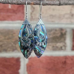 May include: A pair of marquise-shaped dangle earrings featuring iridescent abalone shell inlays set within silver-toned metal frames. The metalwork includes intricate scroll patterns, small circular accents, and a textured leaf design.