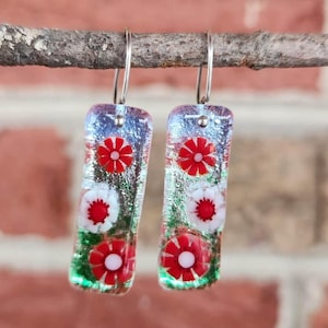 May include: Dangle earrings with a rectangular shape, featuring red and white floral designs. The earrings are made of glass with a dichroic effect, and are suspended from silver-colored hooks. The background is a blurred brick wall.