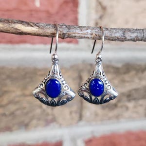 以下が含まれることがあります： 扇形のデザインの銀色のイヤリングのペア。各イヤリングは、華やかな透かし彫りのフレームにセットされた、鮮やかな楕円形の濃い青色の石が特徴です。イヤリングは、シンプルな湾曲したイヤーフックから吊り下げられています。