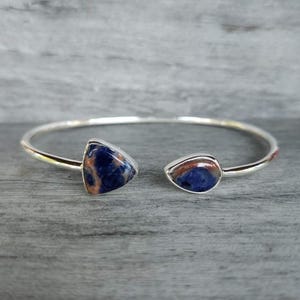 May include: A silver-toned open cuff bracelet featuring two polished, teardrop-shaped stones. The stones display a marbled pattern of deep blue and earthy brown tones. The metal band is smooth, thin, and circular, designed to be worn around the wrist.