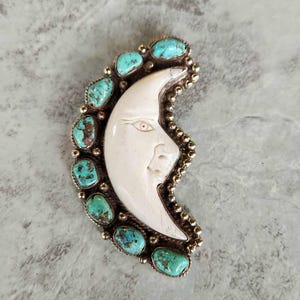 May include: A crescent moon pendant with a carved face, surrounded by turquoise stones and a silver-tone metal border. The moon is white, and the turquoise stones are a vibrant blue-green.