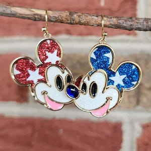 K&ouml;nnte beinhalten: Paar Ohrringe in Mickey Mouse-Form. Einer hat roten Glitzer mit wei&szlig;en Sternen, der andere blauen Glitzer mit wei&szlig;en Sternen. Beide haben ein wei&szlig;es Gesicht, schwarze Augen, einen rosa Mund und einen goldenen Rand.