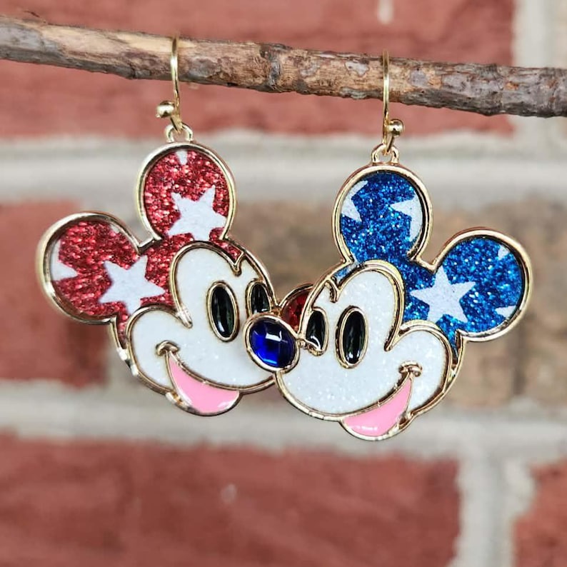 K&ouml;nnte beinhalten: Paar Ohrringe in Mickey Mouse-Form. Einer hat roten Glitzer mit wei&szlig;en Sternen, der andere blauen Glitzer mit wei&szlig;en Sternen. Beide haben ein wei&szlig;es Gesicht, schwarze Augen, einen rosa Mund und einen goldenen Rand.