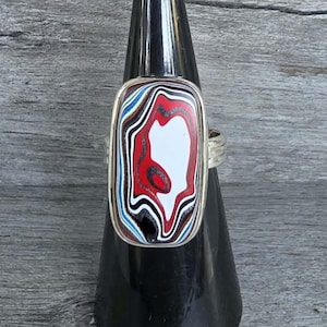 May include: A rectangular ring featuring a polished metal setting and a multi-layered inlay of swirling red, white, black, and blue patterns. The stone has a smooth, glossy surface with distinct, wavy, and organic lines throughout the design.