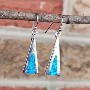 May include: A pair of silver-tone dangle earrings. Each earring features a triangular shape with a vibrant blue opal inlay. The earrings are suspended from a simple hook. The background is a blurred brick wall.