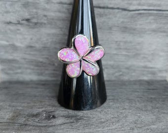 May include: A silver-toned ring featuring a flower design with five petals. The petals are filled with a pink, opalescent material. The ring is displayed on a black, cone-shaped stand against a gray wooden background.