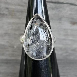 May include: A teardrop-shaped ring featuring a clear gemstone with visible black needle-like inclusions. The stone is set in a smooth, polished silver-toned metal bezel, showcasing a natural, translucent appearance with dark, linear patterns throughout.