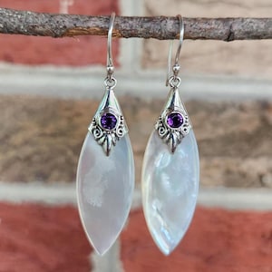 May include: A pair of drop earrings featuring elongated, teardrop-shaped white mother-of-pearl pendants. Each earring is topped with a small, round, deep purple amethyst gemstone set in a decorative silver-toned metal cap with a hook wire.