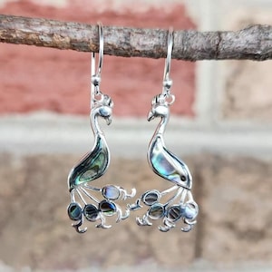 May include: Silver peacock earrings with iridescent abalone shell accents. The earrings feature a detailed peacock design with a crown and a fan-shaped tail. The earrings are hanging from a wooden branch against a blurred brick background.