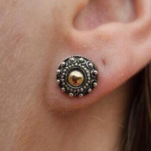 May include: A silver-toned stud earring with a gold-colored center, featuring a detailed, circular design. The earring is secured to an ear, showcasing its intricate, vintage-inspired aesthetic. The design includes small, raised circular details around the gold center.