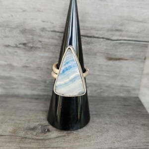 May include: A silver ring with a triangular, blue and white stone. The ring is displayed on a black cone-shaped stand against a light gray wooden background. The stone has a marbled appearance.