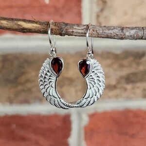 May include: Silver dangle earrings with a wing design. Each earring features a teardrop-shaped, deep red gemstone at the top and a detailed feather pattern. The earrings are displayed against a brick background.