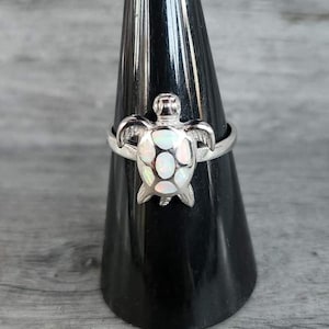 May include: A silver ring featuring a turtle design with a white and iridescent shell. The turtle has detailed flippers and head. The ring is displayed on a black cone-shaped stand against a gray wood-grain background.
