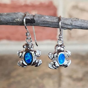 May include: Silver-toned frog earrings with a blue opal center. The earrings are detailed with textured legs and arms, and a simple hook closure. The frog design is a whimsical and unique jewelry piece.