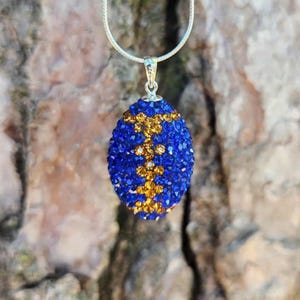 May include: A blue and gold crystal pendant necklace. The oval-shaped pendant is covered in small, sparkling blue crystals with a gold floral design. The necklace has a silver chain and clasp, perfect for adding a touch of elegance to any outfit.
