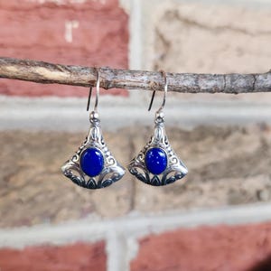 以下が含まれることがあります： 扇形のデザインの銀色のイヤリングのペア。各イヤリングの中央には、精巧なフィリグリーのディテールに囲まれた、大きくて丸い濃い青色の石があります。イヤリングはシンプルなフックから吊り下げられています。