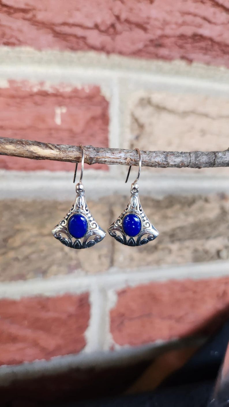 以下が含まれることがあります： 扇形のデザインの銀色のイヤリングのペア。各イヤリングの中央には、精巧なフィリグリーのディテールに囲まれた、大きくて丸い濃い青色の石があります。イヤリングはシンプルなフックから吊り下げられています。