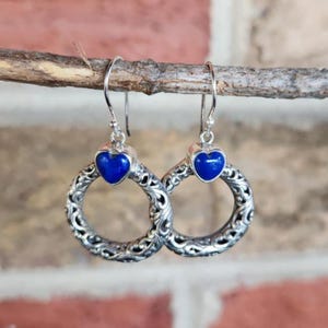 Puede incluir: Pendientes plateados con una piedra azul oscuro en forma de coraz&oacute;n y un dise&ntilde;o de aro calado. Los pendientes cuelgan de un fino gancho de alambre. El fondo es una vista borrosa de una pared de ladrillos y una rama de madera.
