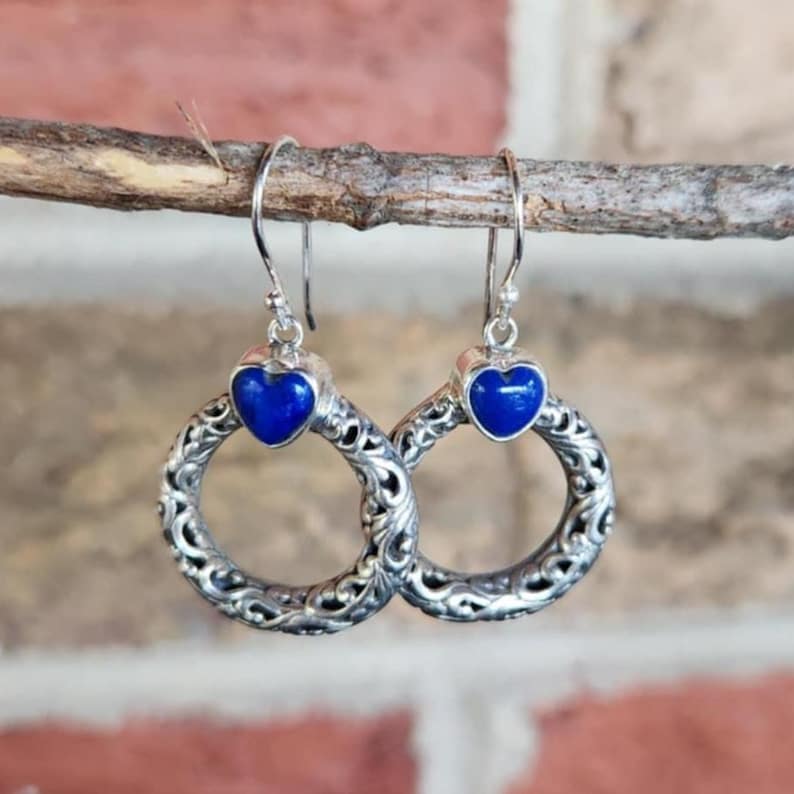 Puede incluir: Pendientes plateados con una piedra azul oscuro en forma de coraz&oacute;n y un dise&ntilde;o de aro calado. Los pendientes cuelgan de un fino gancho de alambre. El fondo es una vista borrosa de una pared de ladrillos y una rama de madera.