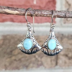 May include: A pair of silver earrings with a light blue oval stone set in a decorative, openwork design. The earrings are suspended from a curved hook and are displayed against a blurred background.